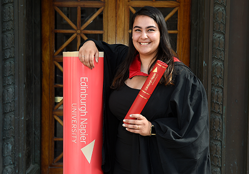 Jade Lindsay wearing graduation gown, standing next to large Edinburgh Napier University scroll prop and holding an Edinburgh Napier University scroll holder.