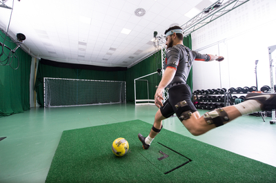 Participant is attached to sensors and monitors to evaluate performance and are getting ready to kick a football into the goal net. 