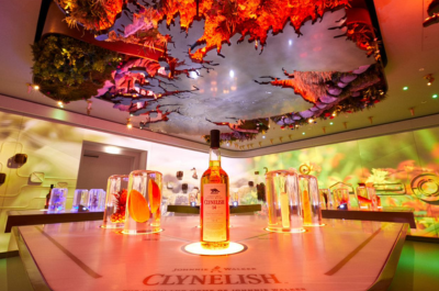 Table set with Clynelish whisky with light surrounding it. The ceiling can be seen with grass and leaves coming out of it above the bottle and glasses with fruit. 