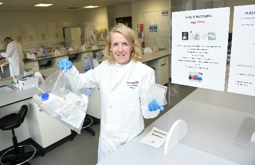 Lisa McMillan, in lab coat and holding a transparent bag, against a backdrop of Sighthill lab