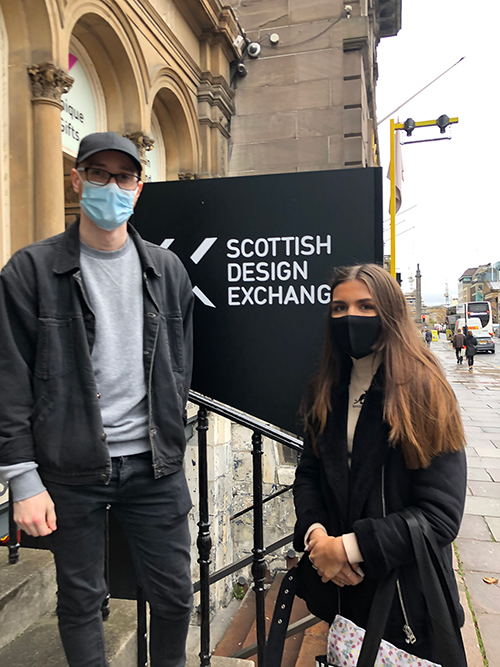 Two Edinburgh Napier students, in masks, stand by sign saying Scottish Design Exchange